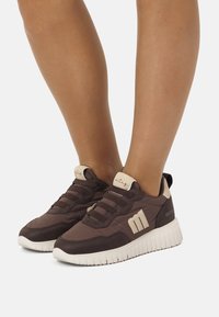 Zapatillas deportivas marrones con parte superior de tela texturizada, acentos crema y suelas de goma blancas. Presentan una puntera redondeada y un diseño con cordones.
