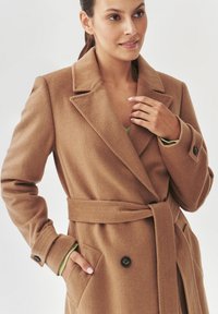 Abrigo de mezcla de lana beige con cintura ceñida por un cinturón, solapas de muesca y botones negros. Presenta bolsillos laterales y una textura suave.