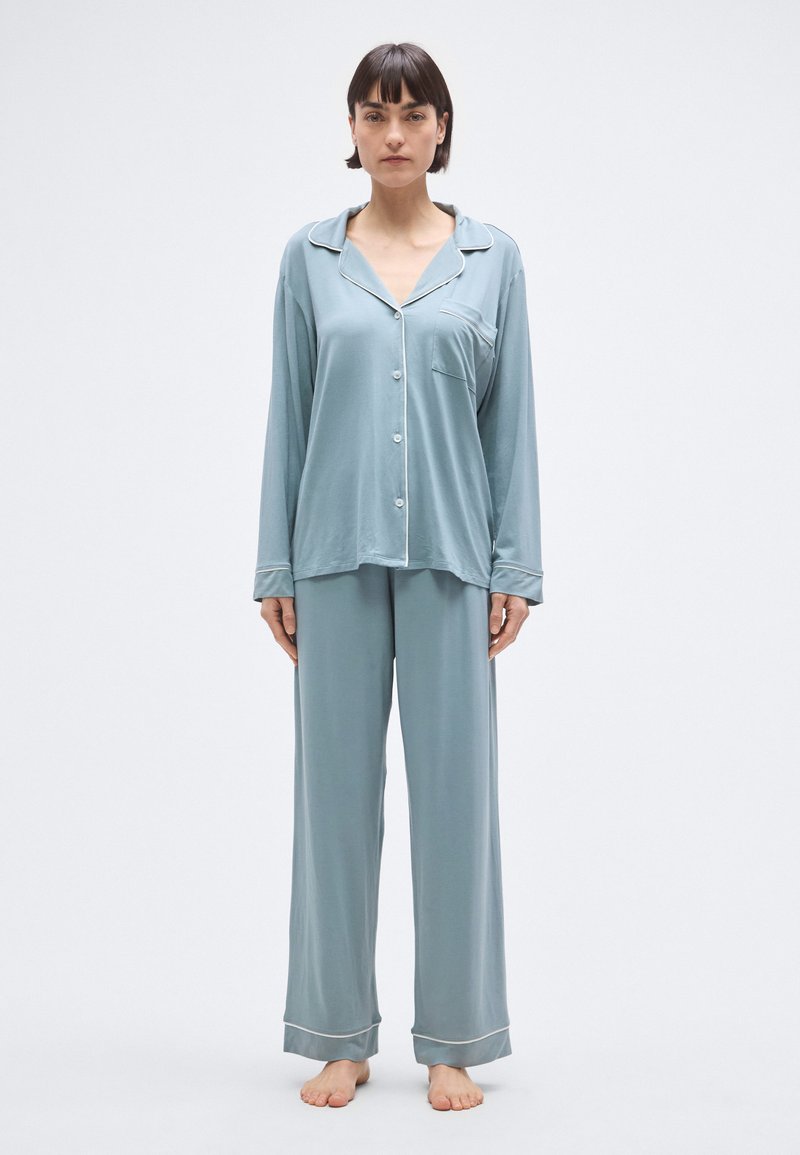 Világoskék pizsamaszett puha anyagból, gombokkal és zsebbel az ingén, a mandzsettán és a nadrágon kontrasztos fehér szegéllyel.