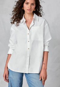 White button-up shirt with a pointed collar, single chest pocket, and rolled-up sleeves. Made of smooth fabric with a relaxed fit.