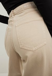Jeans beige a vita alta con una vestibilità su misura, dotati di tasche posteriori e cuciture visibili, realizzati con un tessuto liscio e testurizzato.