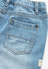 Tissu en denim bleu clair avec une seule poche arrière, une couture orange, un rivet métallique sur le coin de la poche et une partie d'une étiquette en tissu visible.
