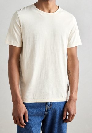 Mężczyzna noszący prosty, białawy T-shirt z krótkim rękawem i subtelnymi paskami oraz niebieskie dżinsy, widoczny tatuaż na lewym przedramieniu.