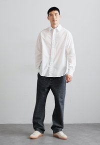 Vit långärmad skjorta med knappar, mörka denimjeans och vita sneakers. Modellen står på ett ljusgrått golv framför en enkel bakgrund.