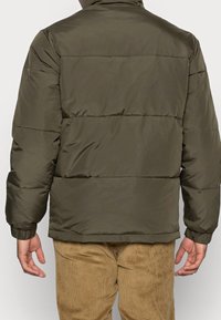 Olivgrön puffjacka med horisontella sömmar, hög krage, elastiska ärmmuddar och en slät, matt yta. Lämplig för kallt väder.