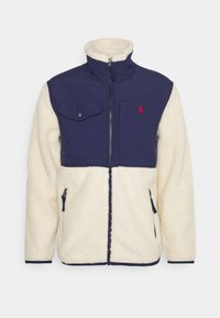 Fleecová bunda s tmavě modrou horní částí a krémovou dolní částí. Má frontální zip, dvě boční kapsy a kapsu na prsou se zápinkou.