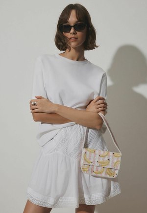 Sieviete ar īsiem brūniem matiem, valkā melnas saulesbrilles, baltu topu un svārkus, rokās tur rozā rokassomu ar dzelteniem banānu rakstiem.