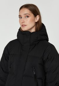 Chaqueta de abrigo negra con cuello alto, con una textura suave, bolsillos laterales con cremallera y un diseño aerodinámico para proporcionar calor.