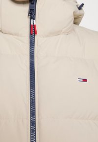 Beige puffjacka med hög krage och marinblå dragkedja, med röda och vita dragkedjehandtag samt en liten broderad logga på bröstet.