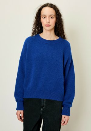 Blauwe pluizige trui met een ronde halslijn, verlaagde schouders en ribgebreide boorden. Gepaard met zwarte jeans, tegen een neutrale achtergrond.