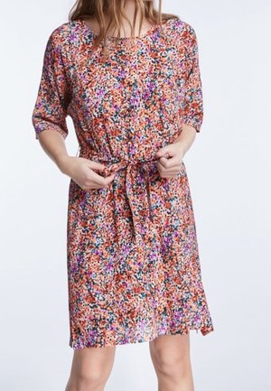 Kobieta regulująca pasek w talii sukienki do kolan w wielokolorowy kwiatowy wzór z rękawami 3/4 na jednolitym białym tle.