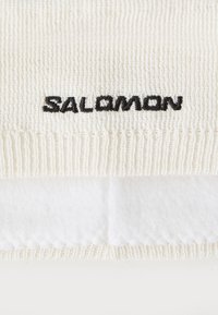 Cappello da sci lavorato a maglia di colore crema con un sottogola in bianco in pile; presenta il logo nero "SALOMON" ricamato sul davanti. Texture morbida, design a coste.