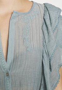 Vaaleansininen paita, jossa on rakenteellinen kangas, hienolla kukka-omenalla ja aallotetulla reunalla. Napitus yksityiskohtana kaula-aukossa.