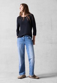Maglietta nera a maniche lunghe, jeans a vita alta azzurri e scarpe beige slip-on; la modella è in piedi contro uno sfondo grigio chiaro, con le mani nelle tasche.