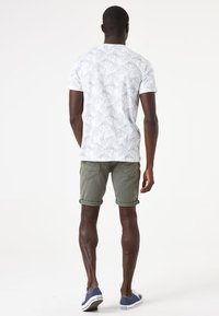 Witte T-shirt met een groen bladpatroon, gecombineerd met opgerolde olijfgroene shorts en blauwe sneakers. Casual ontwerp en lichte stof.