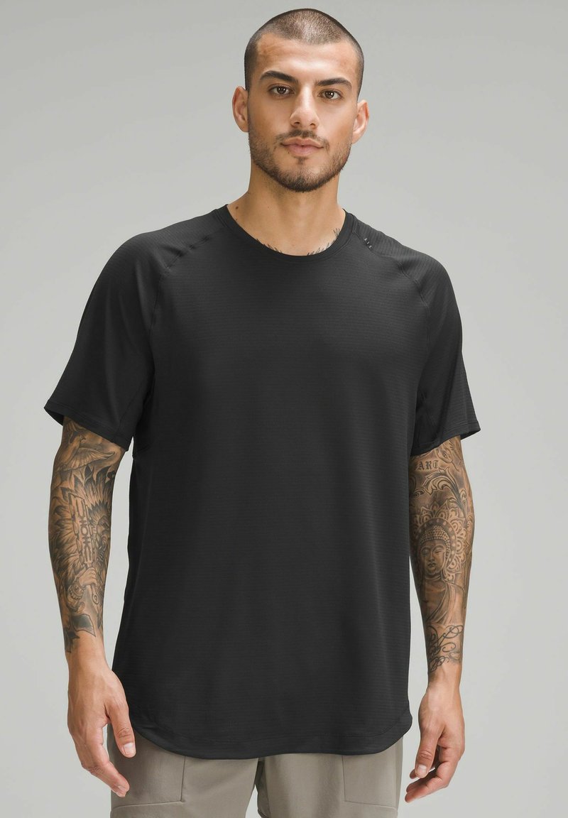 T-shirt de sport noir à manches courtes avec un col rond, en tissu texturé, et manches raglan, porté par un modèle avec des tatouages.