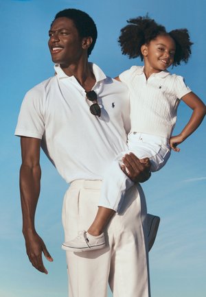 Man in wit poloshirt houdt glimlachend meisje in witte gebreide top en broek tegen een heldere blauwe lucht.