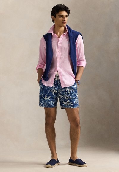 Férfi áll, kezét zsebében tartja, rózsaszín, gombos inget visel, kék mintás rövidnadrágot, tengerészkék pulóvert vállán átvetve, és tengerészkék belebújós cipőt.