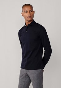 Crna polo majica s dugim rukavima i ovratnikom, s tri gumba na vratu i glatkom, teksturiranom tkaninom. Nosi se s sivoj hlačama.