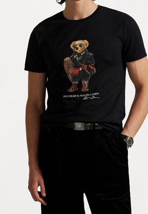 T-shirt noir avec un Polo Bear portant des lunettes de soleil, un équipement d'hiver, tenant une raquette à neige, avec le texte « Polo Bear by Ralph Lauren » en dessous.