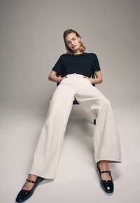 Camiseta de marina, pantalones de tiro alto de pierna ancha color crema y zapatos negros brillantes con tiras en los tobillos. Modelo sentado en una silla contra un fondo neutro.