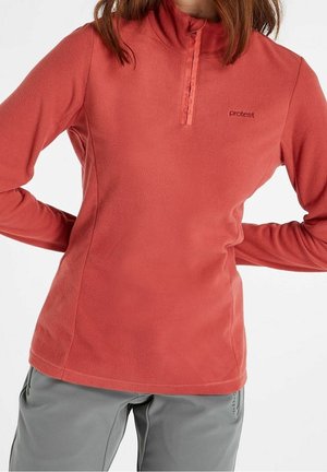 Mujer con un forro polar de color coral con cuello con cremallera lateral y pantalones grises, manos apoyadas en las caderas.