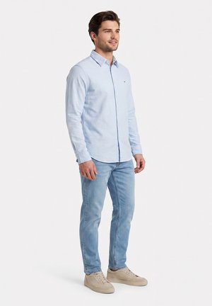 Hombre joven de pie, con camisa de manga larga azul claro, vaqueros azules y zapatillas beige sobre un fondo blanco liso.