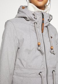 Grey outdoor jacket with a soft white fleece lining, adjustable drawstrings with wooden beads and metal accents, and a zippered front.