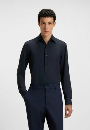 Langärmliges navy-blaues Hemd mit klassischem Kragen und durchgehender Knopfleiste, gefertigt aus glattem Stoff. Kombiniert mit dunklen, maßgeschneiderten Hosen.