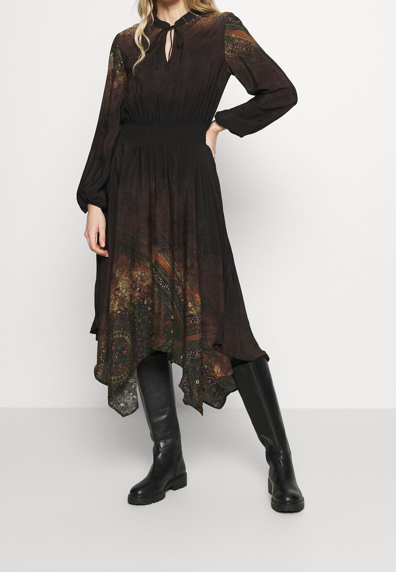 Robe à manches longues marron à motifs, taille cintrée et ourlet asymétrique ; associée à des bottes en cuir noir jusqu'au genou.