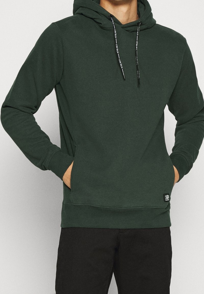 Hombre vestido con una sudadera con capucha de color verde oscuro con cordones negros y las manos en los bolsillos frontales, combinada con pantalones negros.