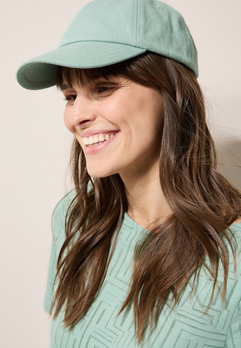 Gorra verde menta hecha de un tejido suave con un visera curva. La persona lleva el pelo largo y ondulado de color castaño y está vestida con una blusa de color menta a juego con un patrón texturizado.