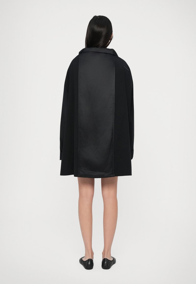 Kvinde iført en oversized sort frakke med et skinnende bagpanel, stående med ryggen til på en ensfarvet hvid baggrund.