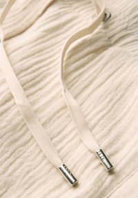 Beige textured fabric with a crinkled design, featuring flat cream shoelaces and metallic tips on both ends.
