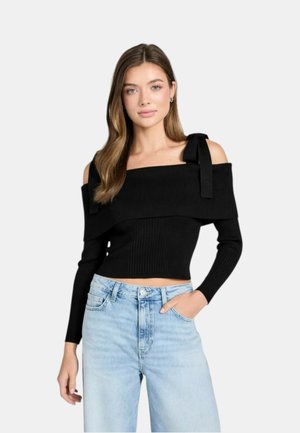 Donna con maglione nero a coste con scollo a barca e dettagli a fiocco, abbinato a jeans azzurri a vita alta, in piedi con una mano in tasca.