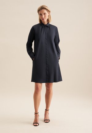 Robe chemise bleu marine à manches longues, fermeture par boutons et poches latérales. Tissu lisse avec une coupe décontractée, arrivant juste au-dessus des genoux.