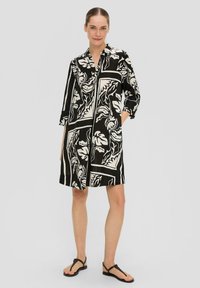 Robe chemise noir et blanc avec des motifs floraux, col classique et manches trois-quarts. Tissu texturé, avec deux poches latérales.