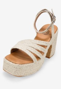 Sandalia de cuña beige con parte superior de tejido trenzado, punta abierta y correa para el tobillo. Textura de yute natural, color neutro y diseño de plataforma resistente.