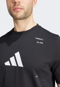 Camiseta de algodón negra con un logo blanco de Adidas, cuello redondo y texto impreso "HANDBALL 60_MIN" en el hombro izquierdo. Textura suave.