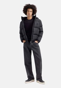 Zwarte pufferjack met een capuchon, voorzien van een ritssluiting en puffy secties, gedragen over een zwart shirt en gecombineerd met losse zwarte jeans en sneakers.