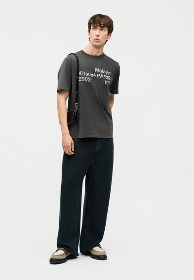 Νεαρός άνδρας που φοράει ξεθωριασμένο μαύρο t-shirt με στάμπα Maison Kitsuné Paris, φαρδιά μαύρη παντελόνια, μπεζ μοκασίνια και μικρή μαύρη τσάντα ώμου.
