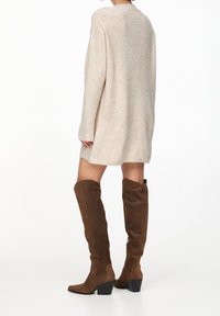 Stivali marroni in suede alti fino al ginocchio con punta quadrata e tacco basso, abbinati a un maglione leggero di colore beige chiaro e dalla vestibilità ampia.