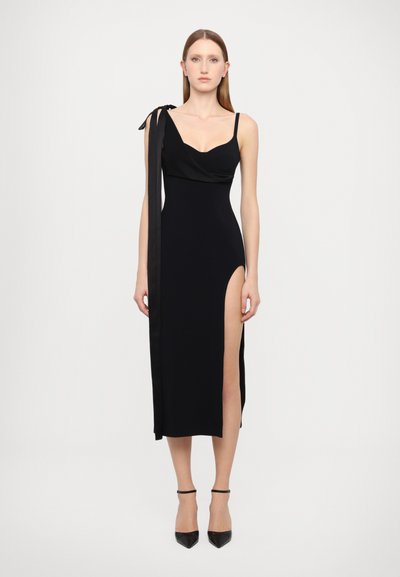 Robe sans manches noire avec un corsage drapé, une bretelle unique attachée en nœud, fente haute sur le côté et silhouette ajustée. Texture lisse.