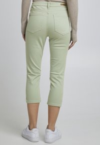 Un pantalon en denim cropped vert clair avec deux poches arrière et des coutures discrètes, associé à des baskets blanches sur un fond neutre.