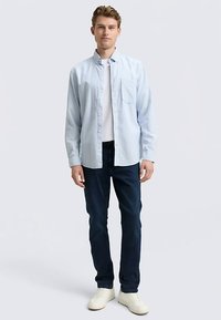 Camicia azzurro chiaro con bottoni e taschino, indossata sopra una t-shirt bianca, abbinata a jeans scuri e sneakers bianche. Tessuto liscio, vestibilità casual.