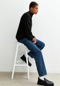 Czarny dziany sweter z wysokim kołnierzem, niebieskie proste dżinsy oraz czarne buty typu slip-on. Model siedzi na białym stołku na tle jednolitej ściany.