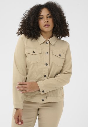 Frau mit lockigem Haar, die eine beige Jacke mit Knöpfen und passende Hose trägt und mit einer Hand auf dem gegenüberliegenden Arm steht.
