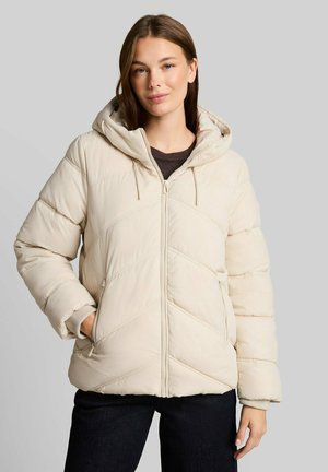 Junge Frau in einer cremefarbenen, gesteppte Daunenjacke mit Kapuze, Hände in den Taschen, vor einem schlichten grauen Hintergrund.