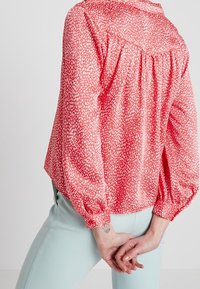 Blusa rossa con motivo a pois bianchi, caratterizzata da un colletto arricciato e maniche lunghe e gonfie. Indossata con pantaloni azzurri chiari.