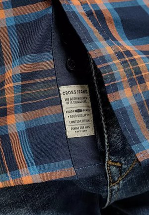 Niebieska i pomarańczowa koszula w kratę częściowo otwarta, odsłaniająca metkę materiałową z napisem "Cross Jeans" oraz pas dżinsów poniżej.